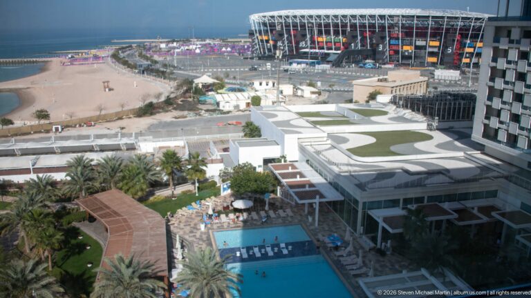 Elevated hotel view across pools, palms, roads, and construction toward Stadium 974 on Doha’s waterfront.