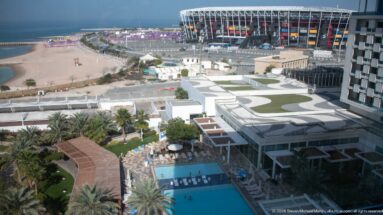 Elevated hotel view across pools, palms, roads, and construction toward Stadium 974 on Doha’s waterfront.