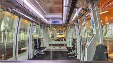 Interior of a glass-walled airport shuttle car with rows of seats and terminal structure visible outside.