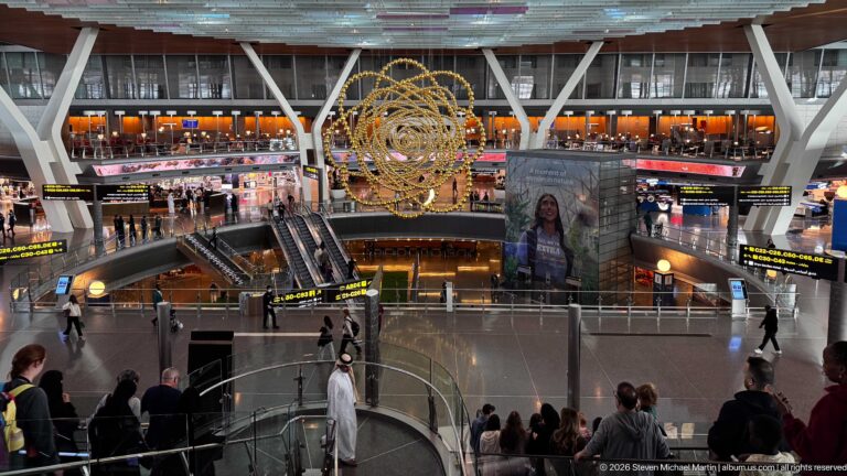 Wide Hamad International Airport concourse with golden sphere sculpture, escalators, shops, passengers, and overhead signs.