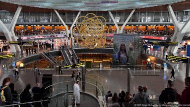 Wide Hamad International Airport concourse with golden sphere sculpture, escalators, shops, passengers, and overhead signs.