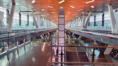 Long multi-level Hamad International Airport concourse with wood-toned ceiling, glass railings, angled columns, and gate signs.