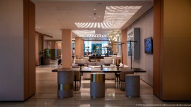 Long modern hotel corridor with seating groups, console tables, cylindrical stools, warm wall panels, and patterned ceiling lighting.