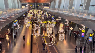 Long airport concourse decorated with glowing crescent moons and lantern shapes above moving walkways and passengers.