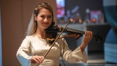 Woman playing violin indoors, holding the instrument beneath her chin with colorful blurred lights behind her.