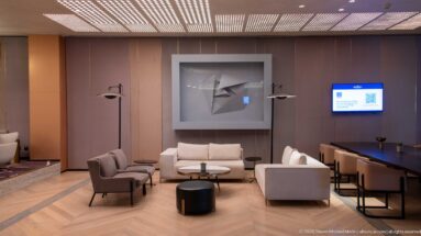 Contemporary hotel lounge with pale sofas, armchairs, black tables, floor lamps, abstract wall art, and a digital display.