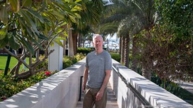 Adult man standing on a shaded walkway bordered by palms, flowering plants, white walls, and dense resort landscaping.