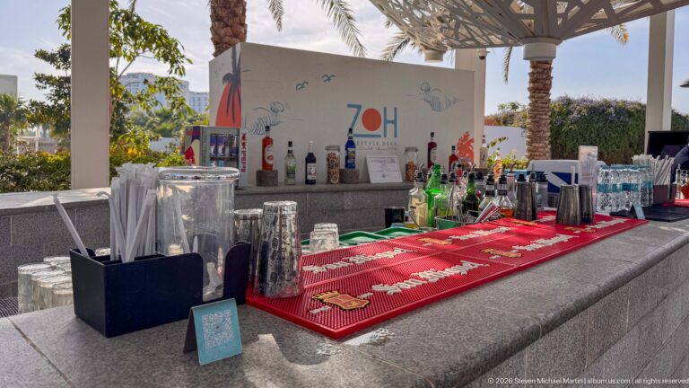 Outdoor resort bar with liquor bottles, glassware, red service mat, and a large 70th-anniversary graphic behind the counter.