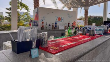 Outdoor resort bar with liquor bottles, glassware, red service mat, and a large 70th-anniversary graphic behind the counter.