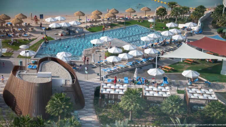 Elevated view of a resort swimming pool, white umbrellas, landscaped lawns, palms, and sandy beach beside Doha Bay.