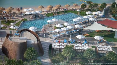 Elevated view of a resort swimming pool, white umbrellas, landscaped lawns, palms, and sandy beach beside Doha Bay.