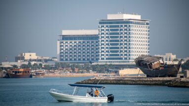 Small motorboat on calm water with a large waterfront hotel and a dark wooden vessel near the shore.
