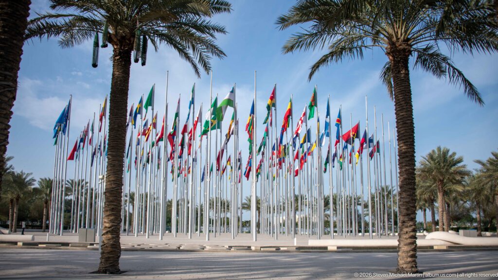 Wide view of Doha’s Flag Plaza with many national flags framed by tall date palms beside a roadway.
