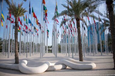 Rows of national flags and palm trees behind white serpentine sculptural seating in Doha’s Flag Plaza.