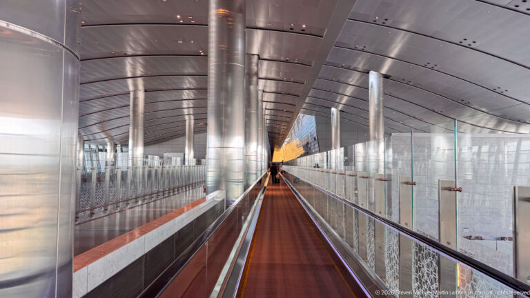 Long moving walkway in a metallic glass-sided corridor with curved ceiling and repeating columns at Hamad International Airport.