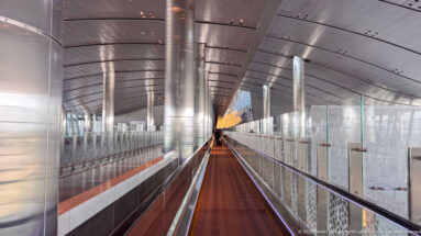 Long moving walkway in a metallic glass-sided corridor with curved ceiling and repeating columns at Hamad International Airport.