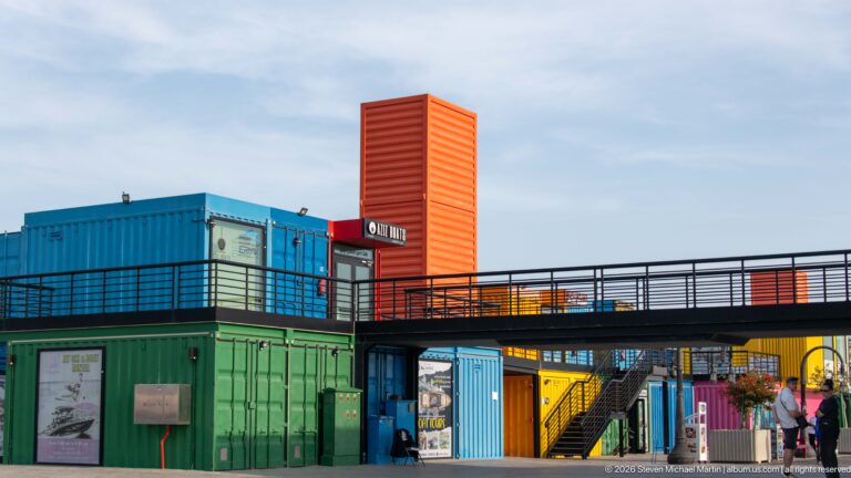 Colorful stacked shipping-container shops linked by a black elevated walkway and staircase at Box Park in Doha.