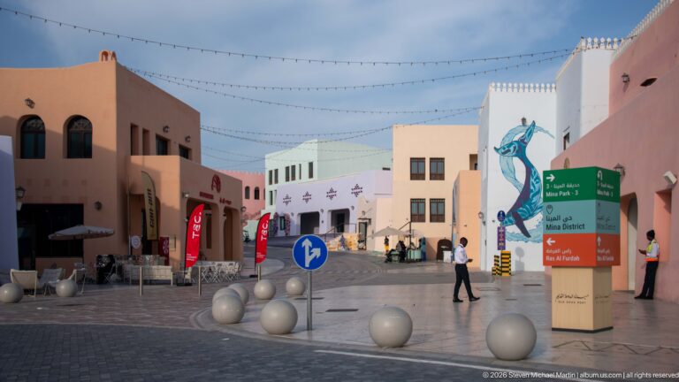 Pastel pedestrian street in Doha’s Mina District with bilingual directional sign, murals, string lights, and round bollards.