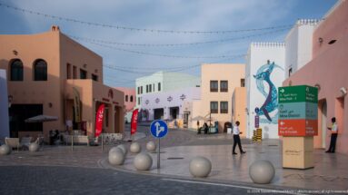 Pastel pedestrian street in Doha’s Mina District with bilingual directional sign, murals, string lights, and round bollards.