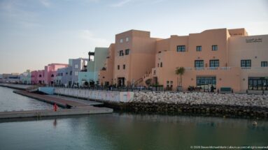 Pastel low-rise buildings lining a calm harbor basin with promenade and rock-edged quay in Doha’s Mina District.