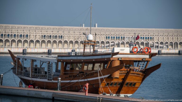 Traditional wooden dhow moored at a quay with raised prow, enclosed passenger deck, mast, and orange life rings.