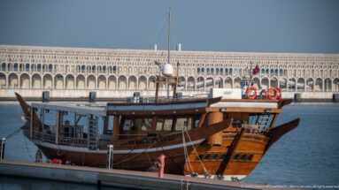 Traditional wooden dhow moored at a quay with raised prow, enclosed passenger deck, mast, and orange life rings.
