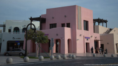 Pink two-story building with rooftop pergolas and decorative trim on a pedestrian street in Doha’s Mina District.