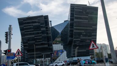 Angular dark-glass office towers beside a busy Doha intersection with cars, pedestrians, traffic lights, and road signs.