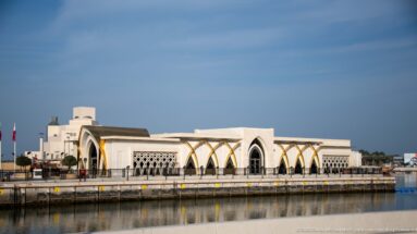 Long pale waterfront building with pointed arches, geometric screens, and gold-toned supports beside a calm harbor basin.