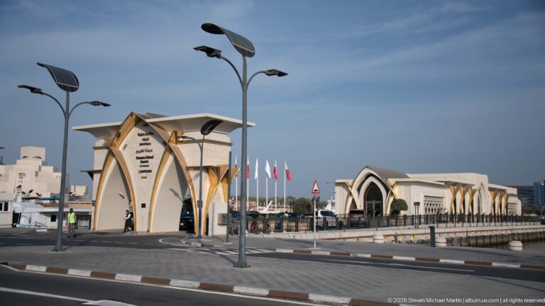 Pointed-arch gateway and pale waterfront buildings with patterned panels and gold-toned supports at Old Doha Port.