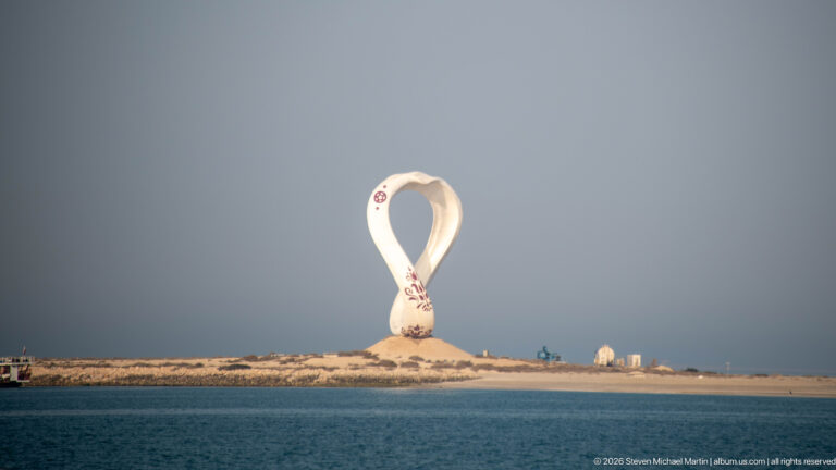 Large white loop-shaped outdoor sculpture standing on a low sandy shoreline beyond calm blue water.