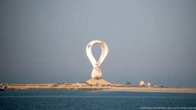 Large white loop-shaped outdoor sculpture standing on a low sandy shoreline beyond calm blue water.