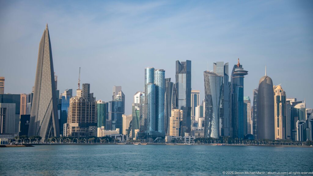 Wide view across Doha Bay toward the West Bay skyline, including a prominent sharply tapered tower at left.