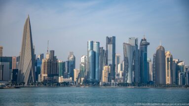 Wide view across Doha Bay toward the West Bay skyline, including a prominent sharply tapered tower at left.