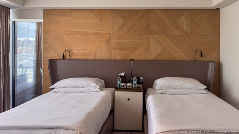 Two single hotel beds beneath a geometric wood-paneled wall with shared bedside cabinet, bottled water, and reading lamps.