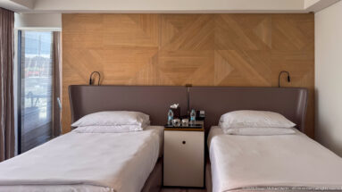 Two single hotel beds beneath a geometric wood-paneled wall with shared bedside cabinet, bottled water, and reading lamps.