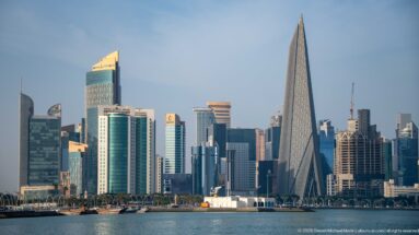 Doha West Bay skyline with a tall tapered triangular tower above traditional wooden vessels along the waterfront.