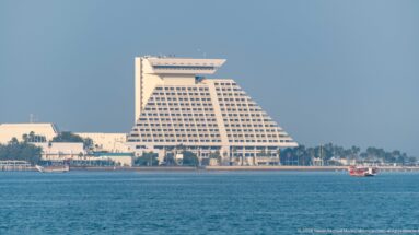 Stepped pyramidal Sheraton Grand Doha hotel viewed across blue water with palms and small boats along the shore.