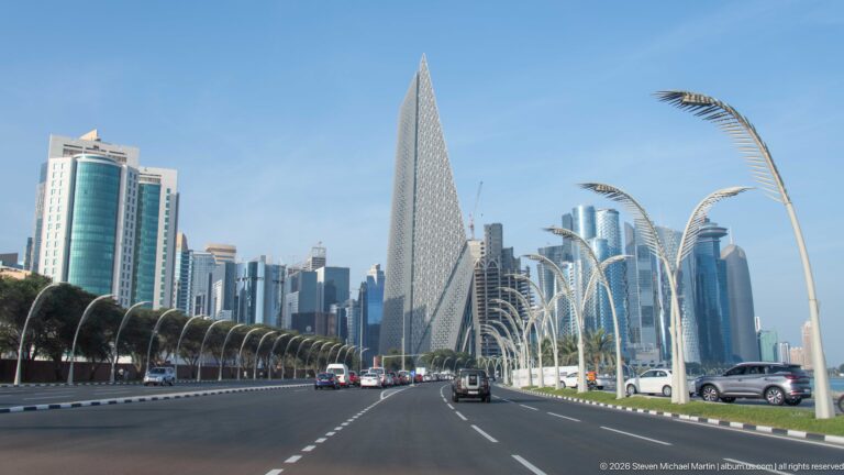 Multilane Doha boulevard leading toward a tall triangular patterned tower and clustered West Bay high-rises.