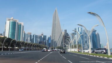 Multilane Doha boulevard leading toward a tall triangular patterned tower and clustered West Bay high-rises.