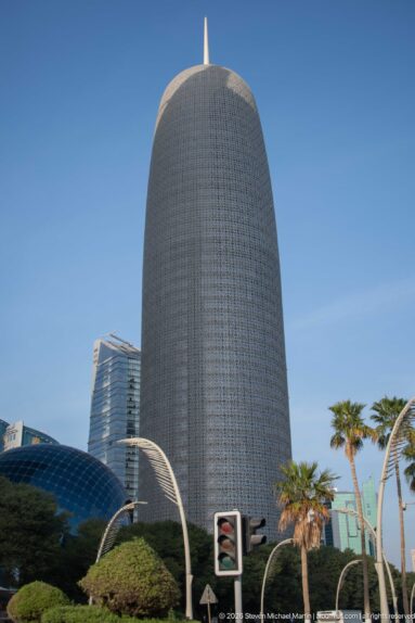 Doha Tower with rounded form, patterned external screen, pointed roof finial, and landscaped street foreground.