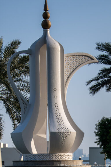 Monumental white dallah coffee-pot sculpture with curved handle, long spout, patterned surface, and palm trees.