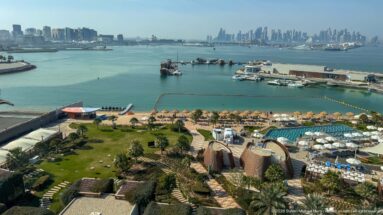 Elevated panorama of resort gardens, pools, beach umbrellas, Doha Bay, boats, and the distant city skyline.