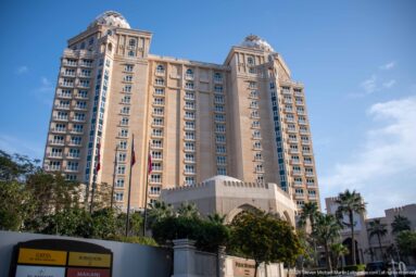 Beige high-rise hotel with regular window bays, arched entrance, landscaping, and Qatari flags.