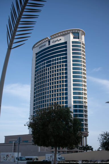 Hilton-branded high-rise hotel with blue-glass side façades and regular window bays under a clear blue sky.