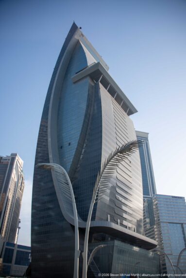 Low-angle view of a sculptural dark-glass high-rise with curved façades, horizontal fins, and a projecting roof.