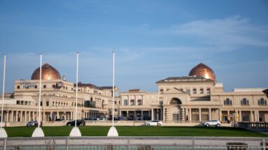 Large cream waterfront complex with two copper-colored domes, central arch, formal lawn, and fountain.