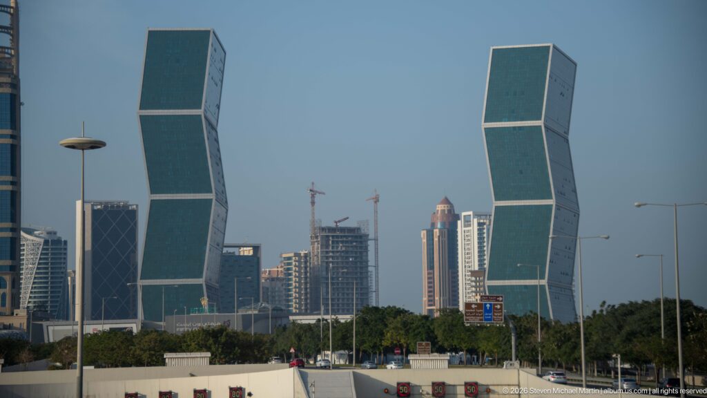Two matching green-glass towers made of offset rectangular blocks with other high-rises and cranes behind.