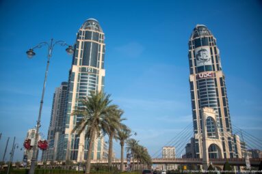 Twin dark-glass high-rise towers with rounded crowns beside cable-supported roadway infrastructure and palms.