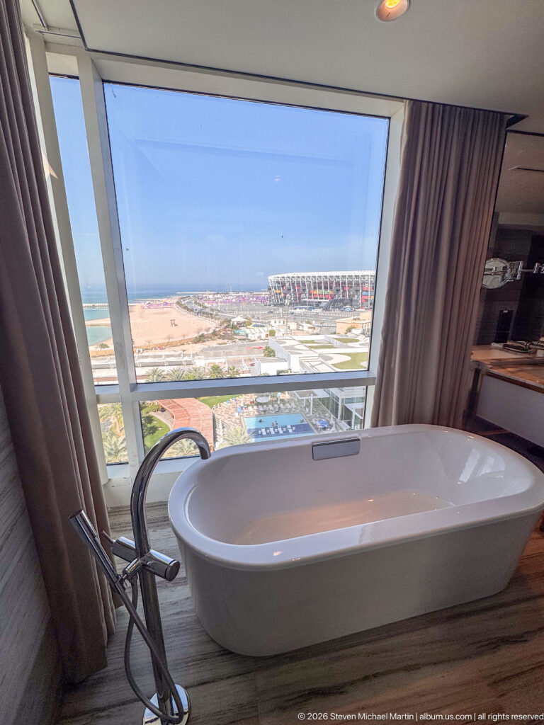 Freestanding white bathtub beside a floor-to-ceiling window, with a stadium and coastal development visible outside.